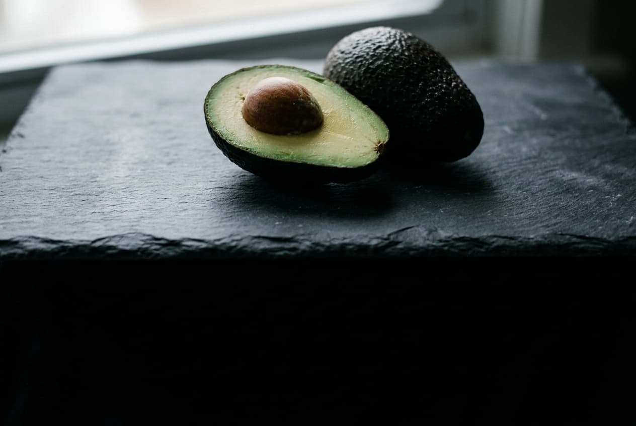 A halved Hass avocado and a whole one on a dark stone slab, lit from a window to the left, the lower half of the frame in shadow